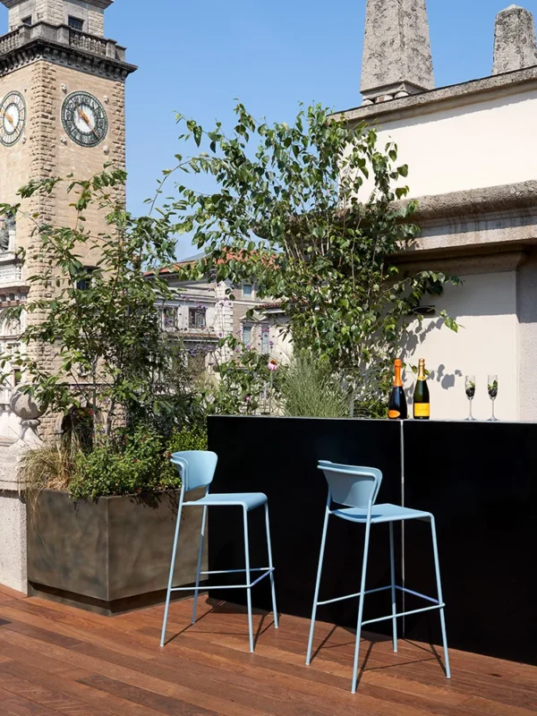 Δίχρωμα σκαμπό μπαρ Lisa Technopolymer με μπράτσα, σε εξωτερικό χώρο με φόντο πράσινο