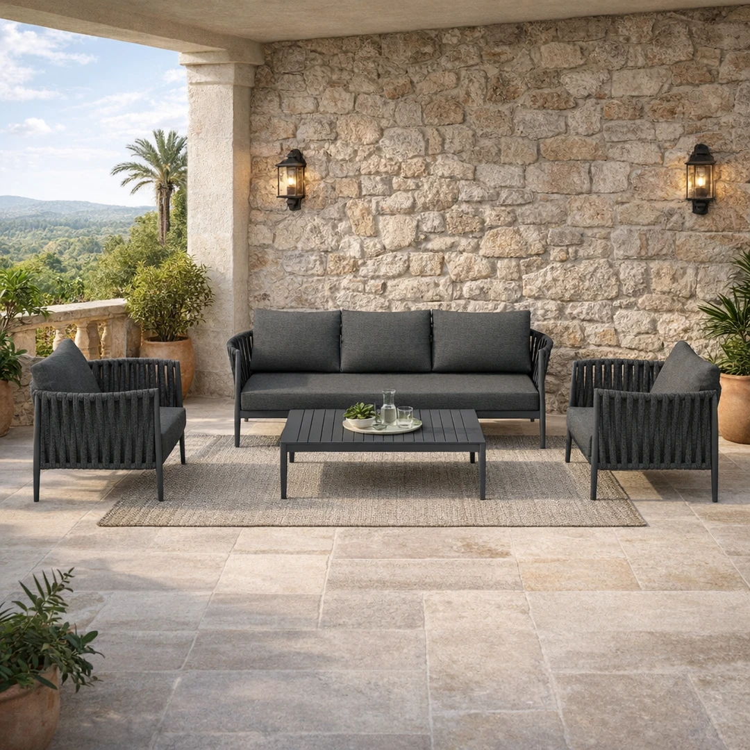 «Σετ επίπλων εξωτερικού χώρου με γκρι καναπέ, δύο πολυθρόνες και τραπεζάκι σε βεράντα βίλας μπροστά από πετρόχτιστο τοίχο.»